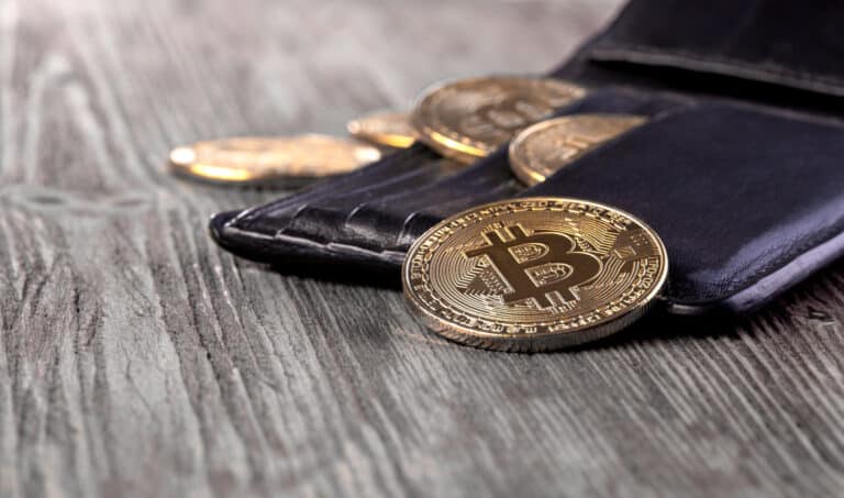 Portefeuille en cuir noir contenant plusieurs pièces physiques de Bitcoin posées sur une table en bois.