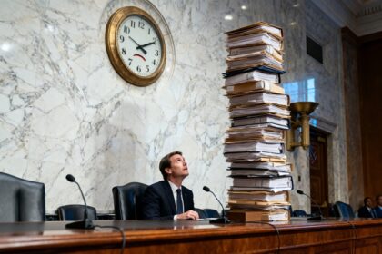 Un homme en costume assis à un pupitre d'audition lève les yeux vers une horloge murale, à côté d'une haute pile de dossiers