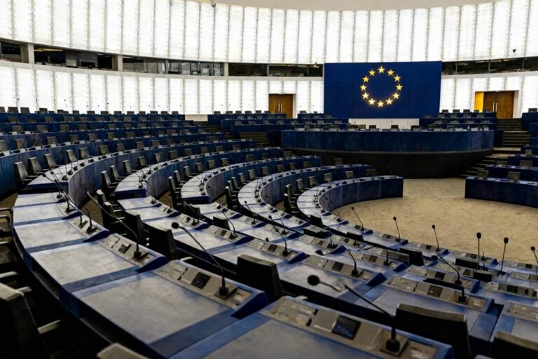 Hémicycle institutionnel européen vide, rangées de pupitres bleus et micros, cercle d'étoiles dorées de l'UE au fond