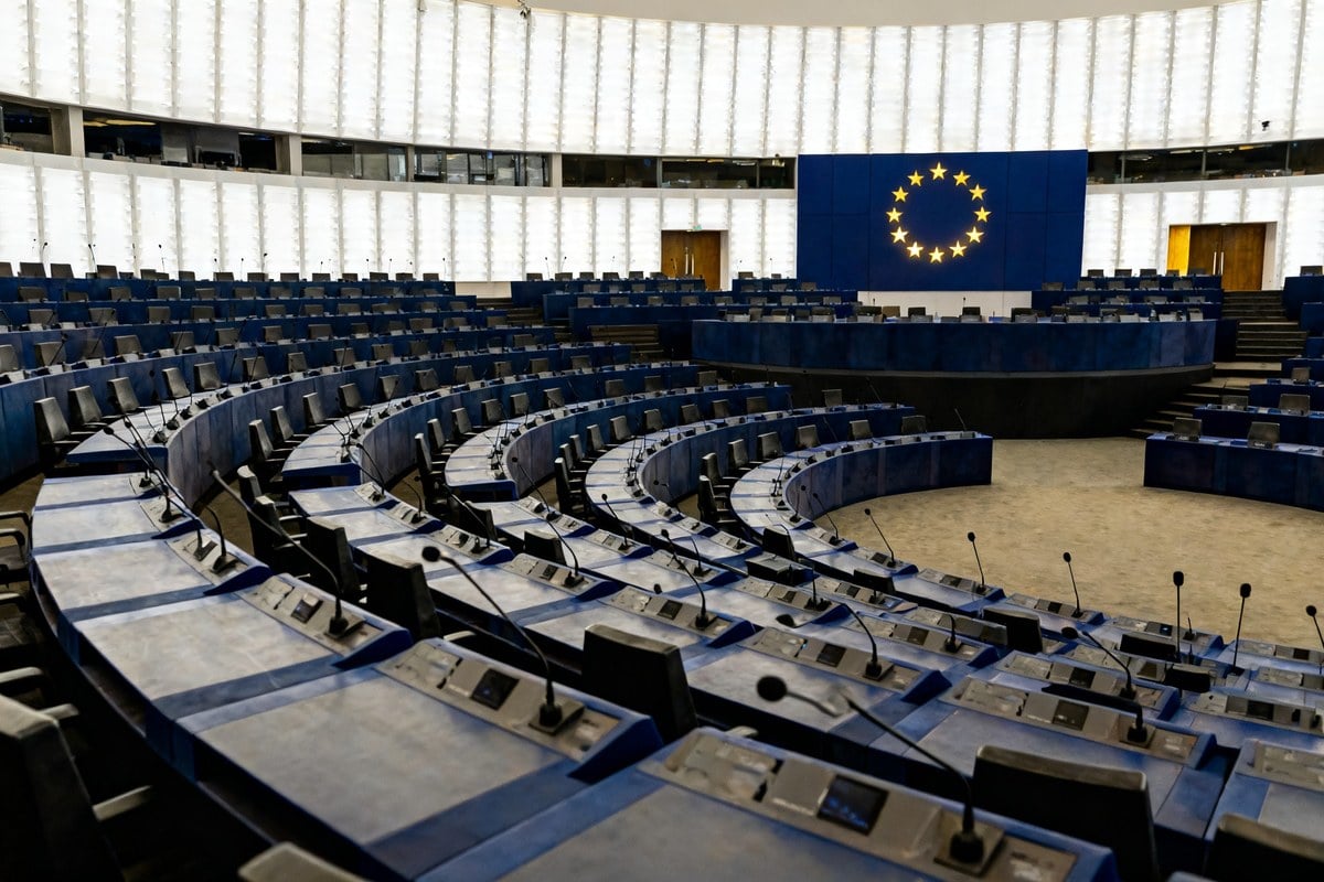 Hémicycle institutionnel européen vide, rangées de pupitres bleus et micros, cercle d'étoiles dorées de l'UE au fond