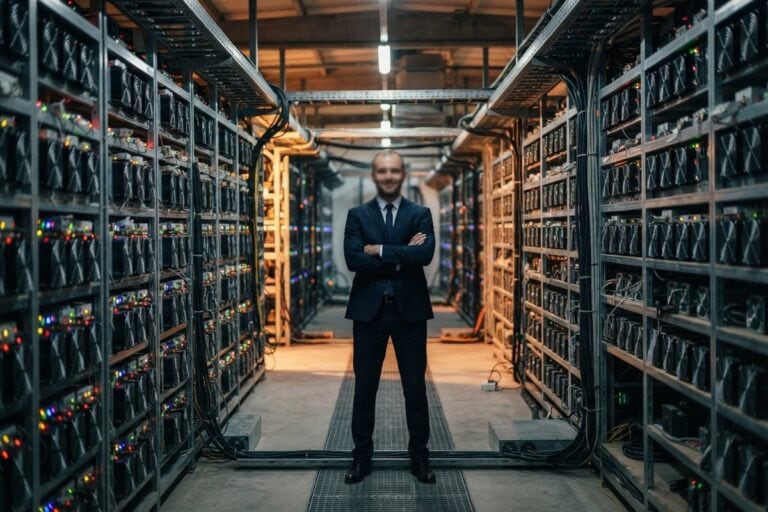 Un homme en costume, bras croisés, au centre d'une allée bordée de machines de minage aux voyants colorés.