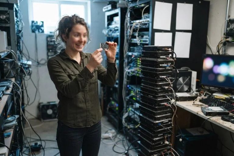 Une femme tient une petite carte électronique face à une haute pile de disques durs dans une salle serveurs